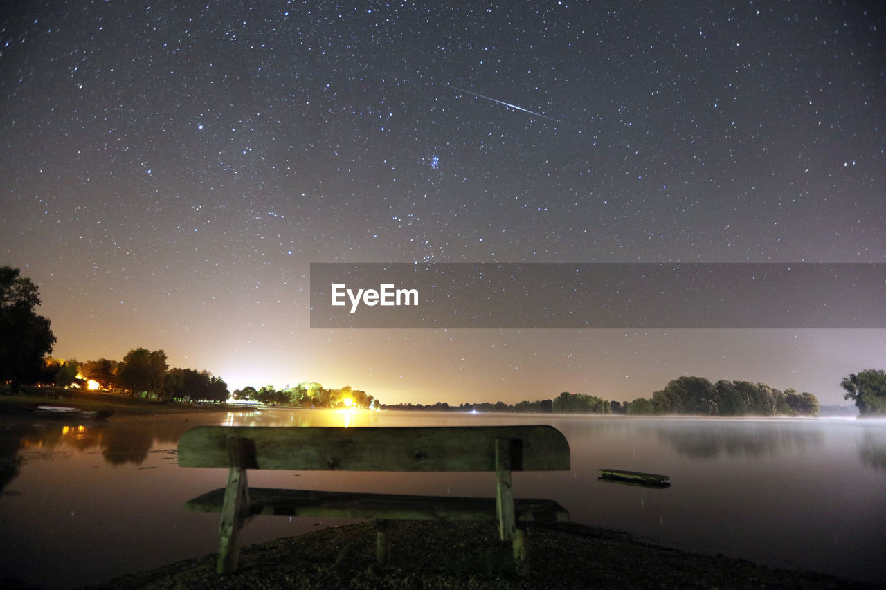 Empty park bench by lake against star field at night
