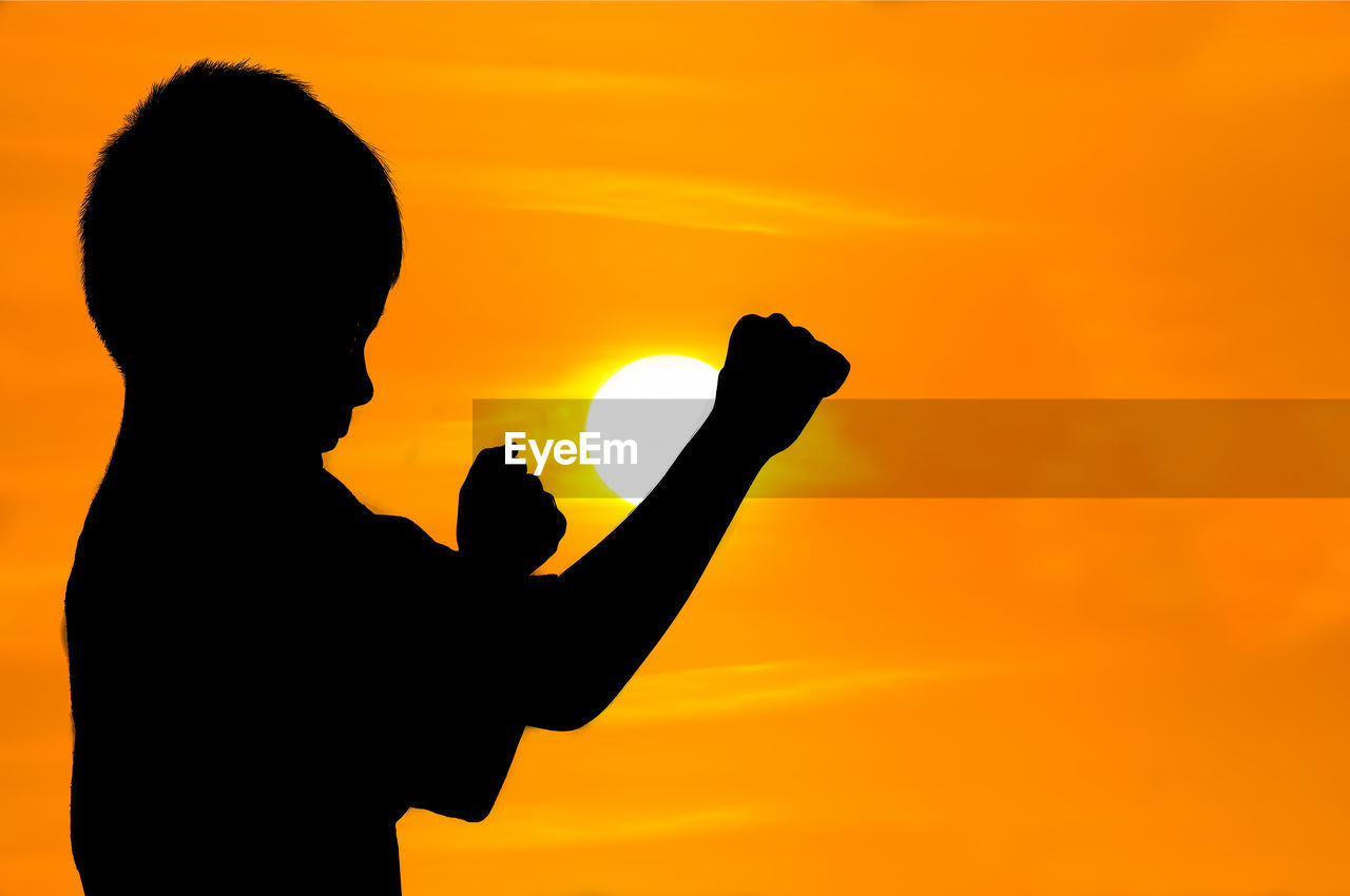 SILHOUETTE BOY HOLDING LEAF AGAINST ORANGE SKY