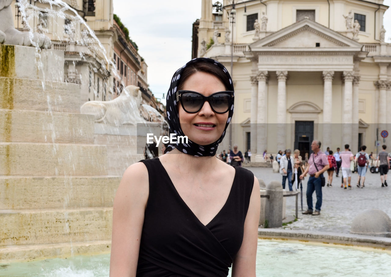 Portrait of smiling woman wearing sunglasses against fountain in town