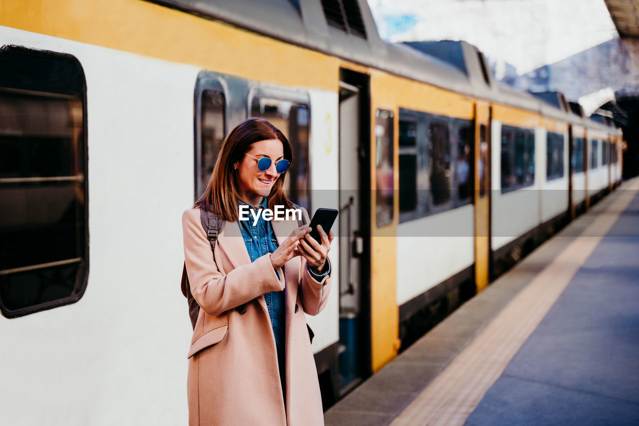 Happy backpacker caucasian woman at platform on train station using mobile phone. travel concept