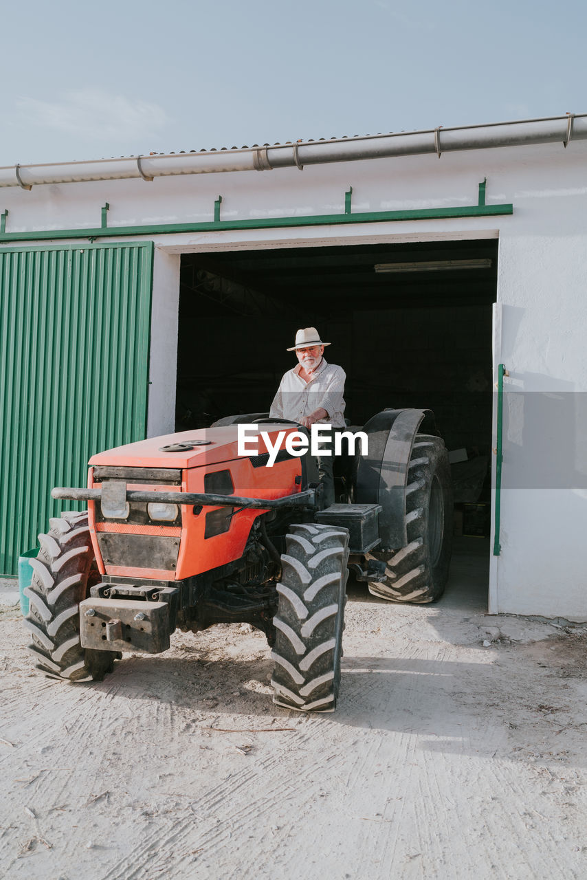 Male farmer in hat driving out of garage in industrial tractor while working in rural area on sunny summer day