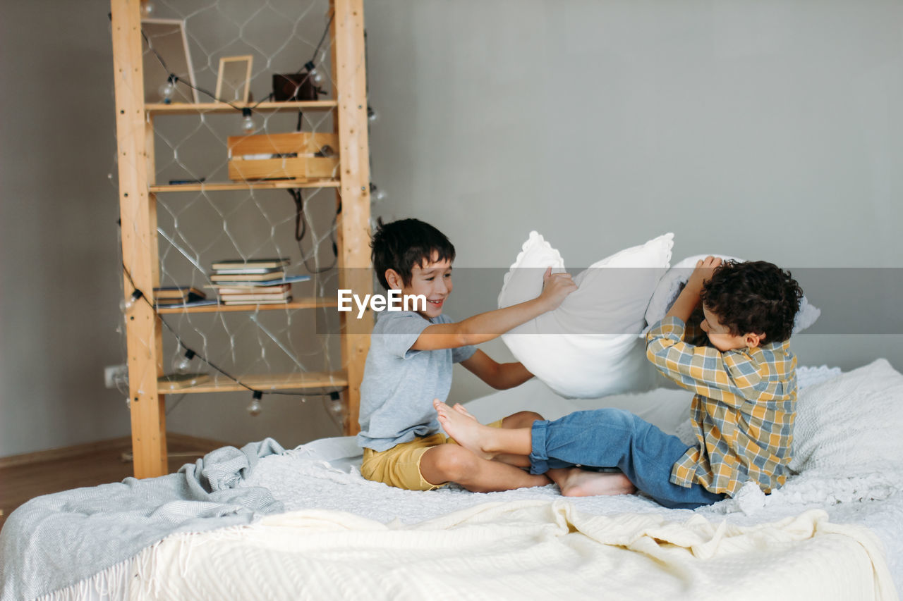 Boys fighting with pillow while sitting on bed