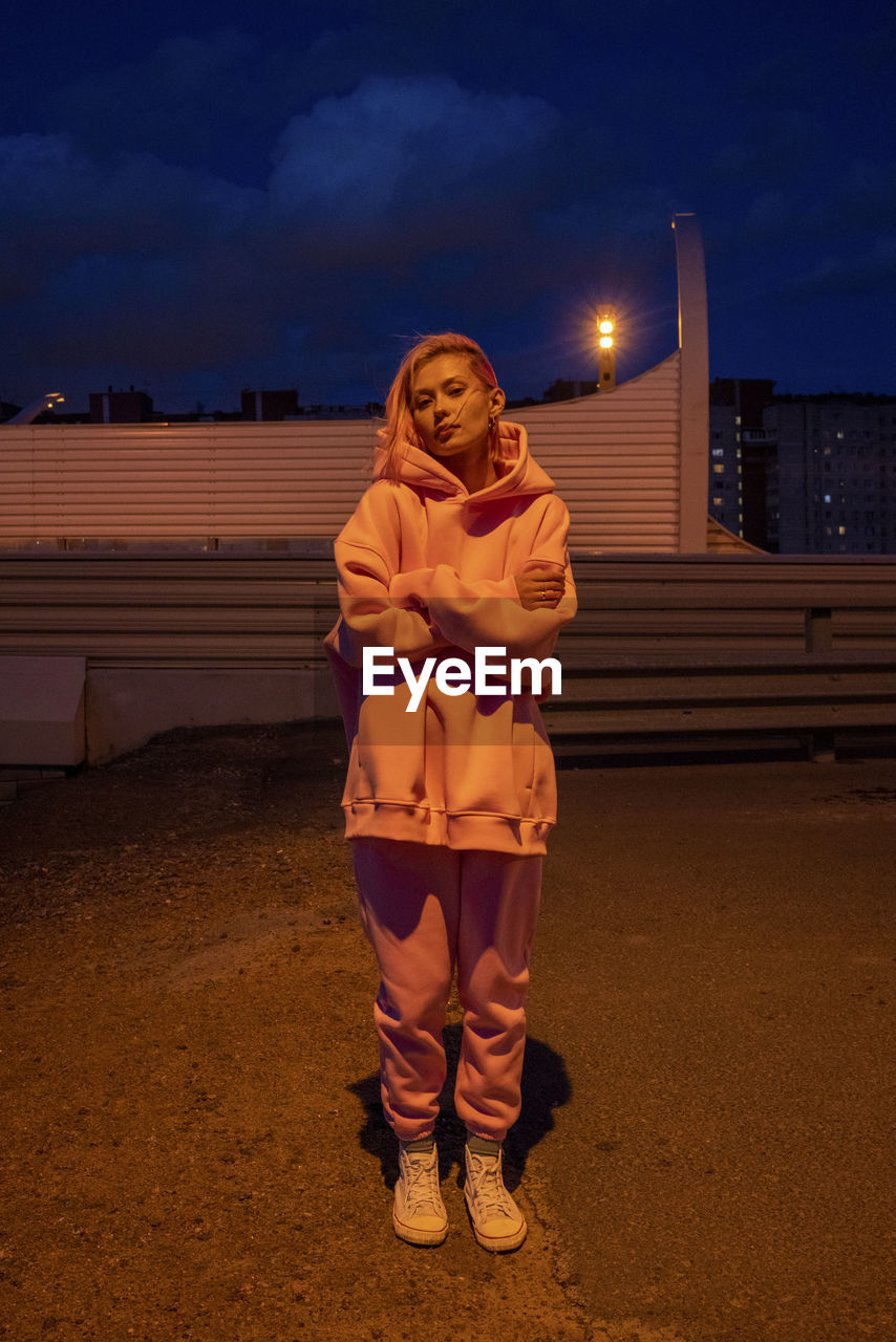 Portrait of young woman with pink hair wearing track suit standing in front of building at night