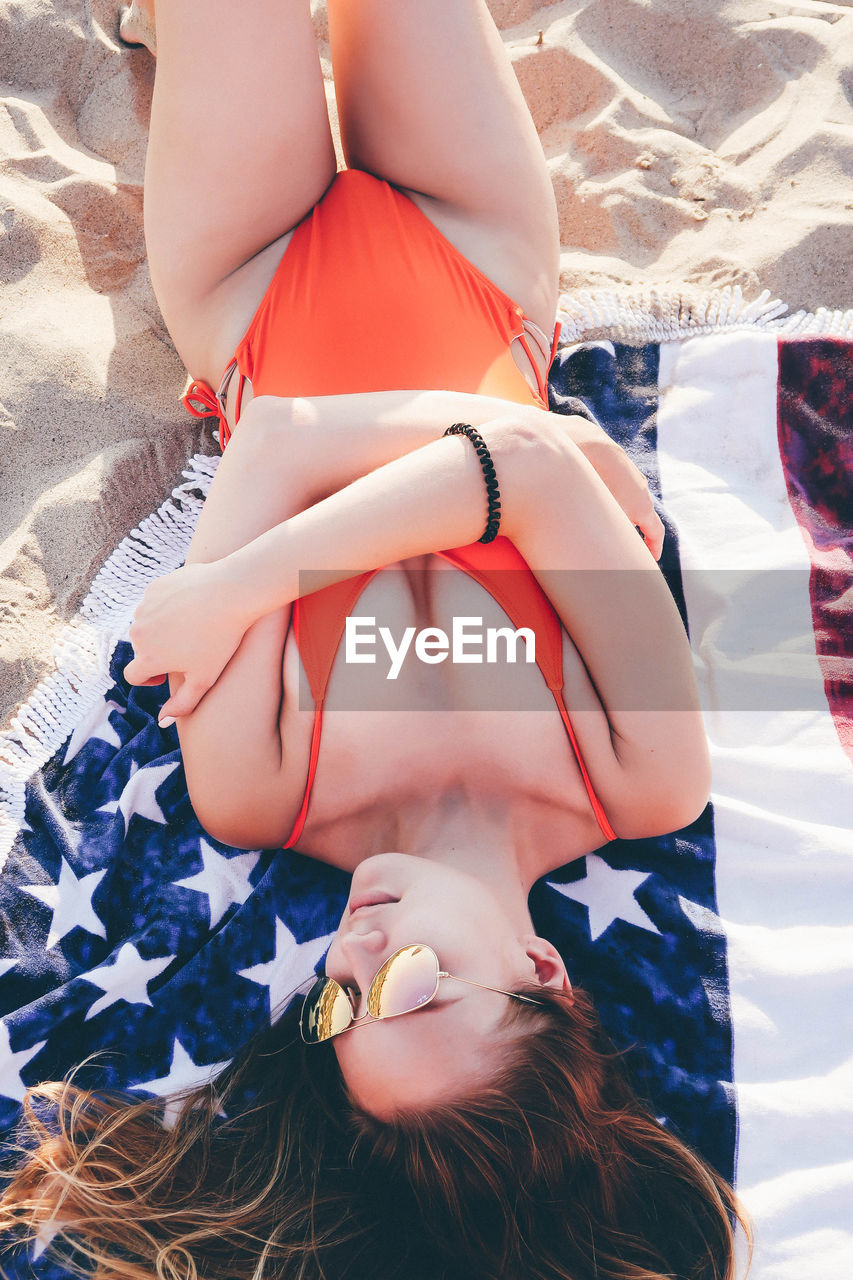 High angle view of young woman wearing sunglasses while lying on blanket at beach