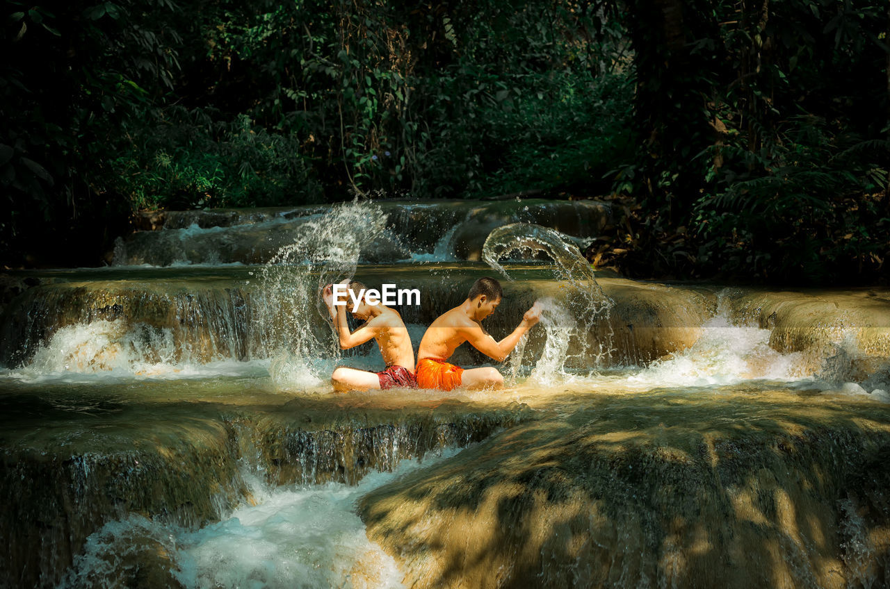 Side view of shirtless male friends splashing water in river
