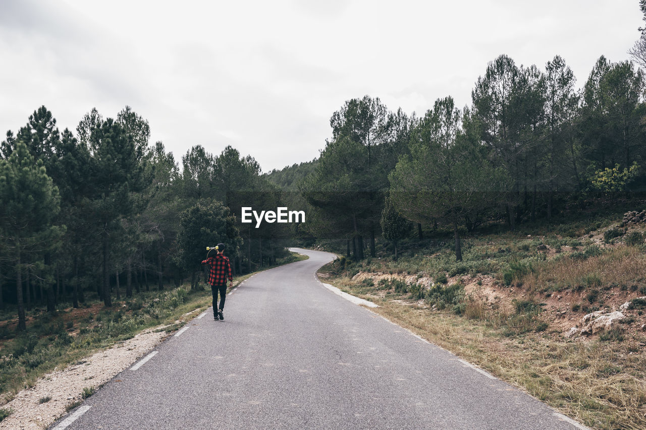 Rear view of man walking on road amidst trees against sky