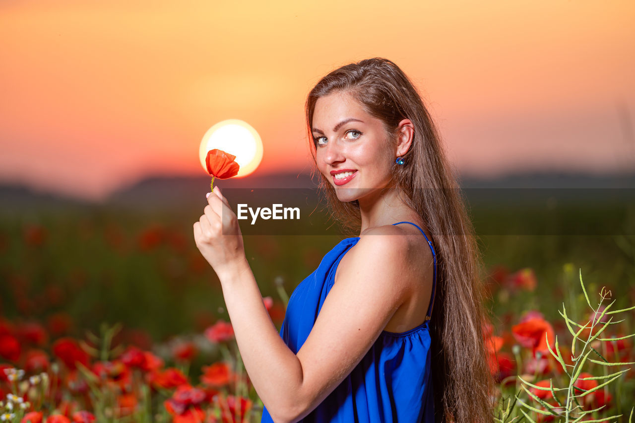 women, one person, plant, smiling, nature, adult, sunset, red, happiness, sky, young adult, beauty in nature, field, landscape, flower, rural scene, emotion, land, flowering plant, summer, female, portrait, long hair, child, hairstyle, lifestyles, cheerful, holding, environment, person, agriculture, meadow, springtime, grass, teenager, sun, positive emotion, freshness, outdoors, sunlight, relaxation, fashion, food and drink, dress, multi colored, plain, wellbeing, enjoyment, waist up, food, clothing, crop, orange color, standing, childhood, side view, vibrant color, brown hair