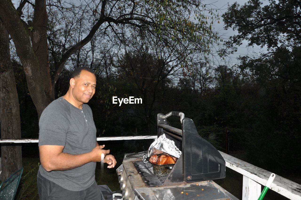 Portrait of mature man preparing food on barbecue grill against trees at dusk