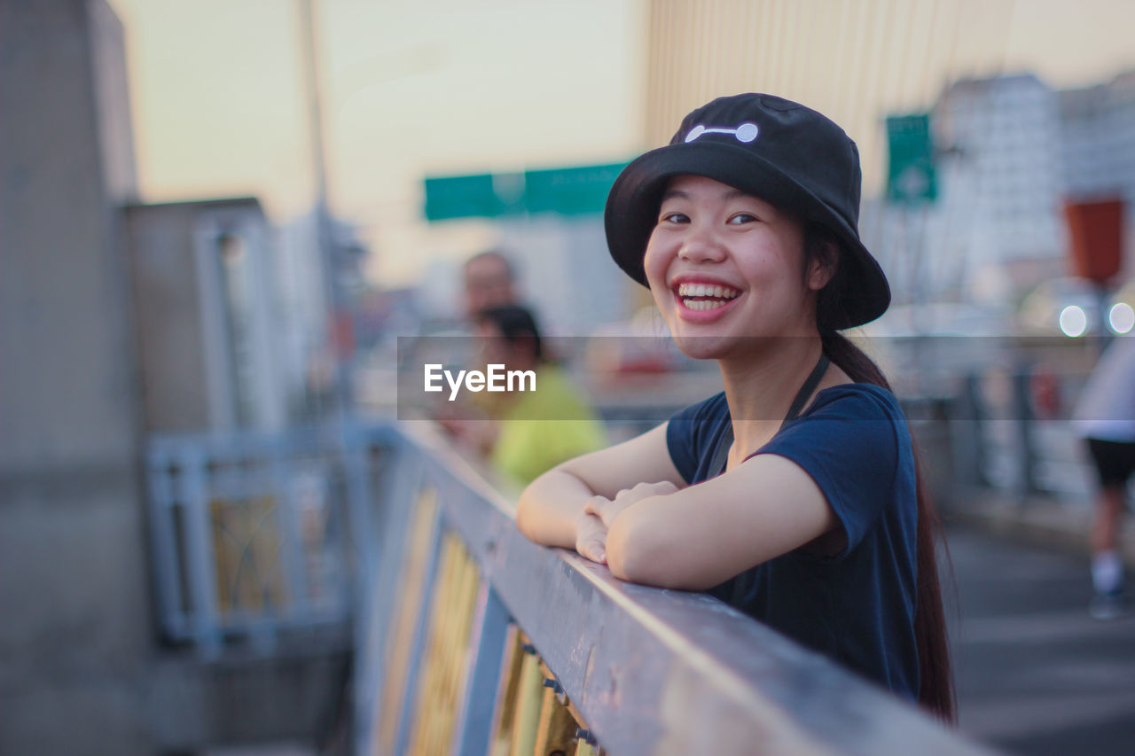 Portrait of smiling young woman wearing hat standing by railing during sunset