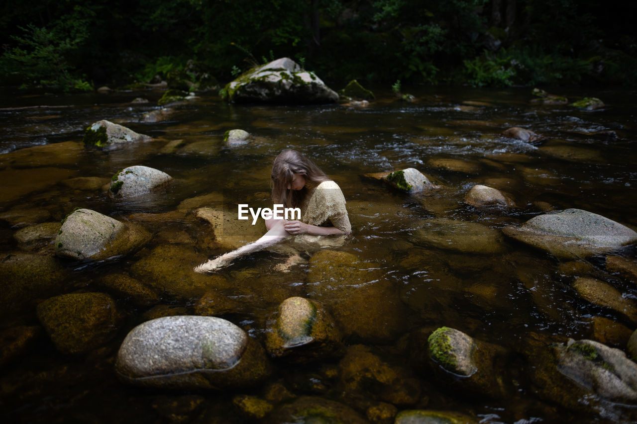 Woman with hair in front of her face sitting in a river between stones wearing a lace dress