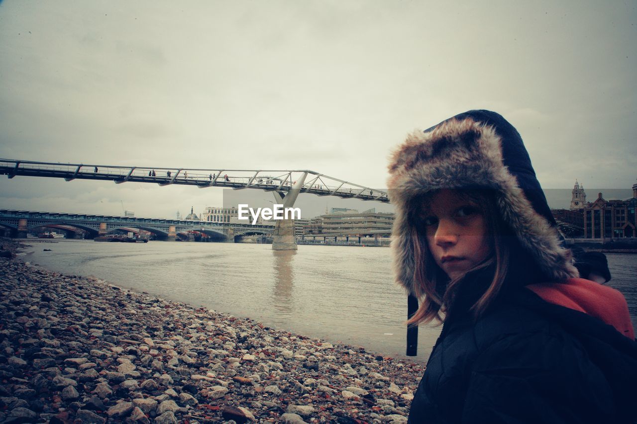 Portrait of girl standing on riverbank against bridge in city