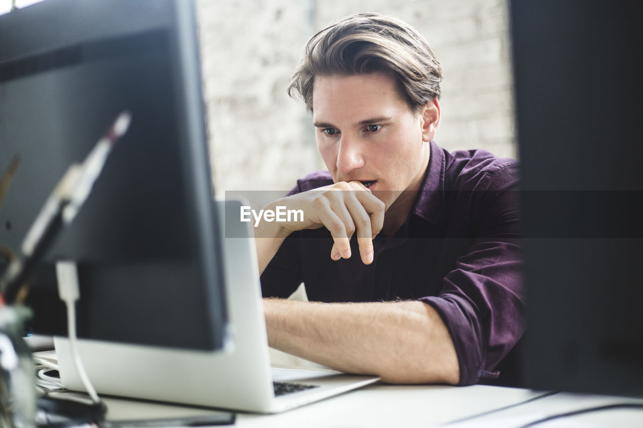 Serious male it professional thinking while coding in laptop at creative workplace