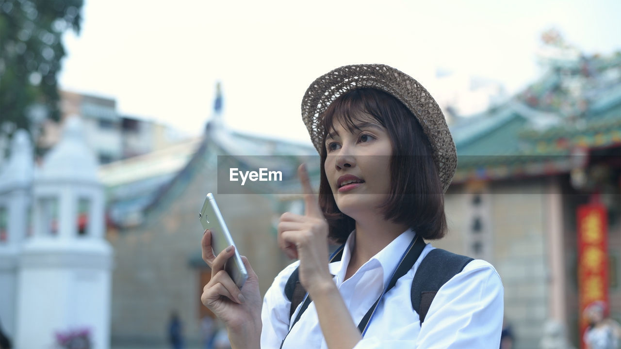 Woman looking away while holding mobile phone in city