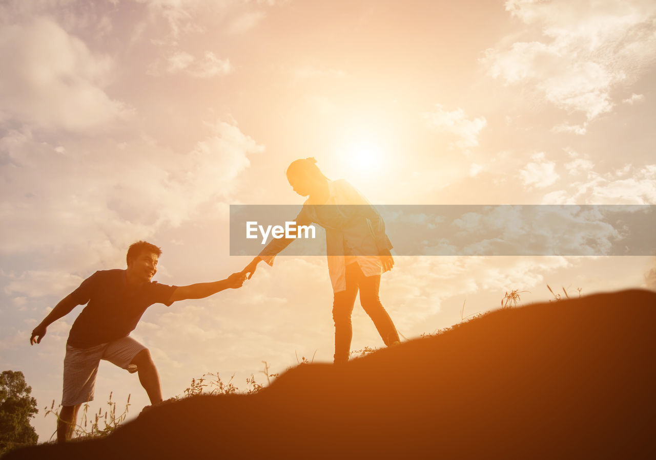 Low angle view of couple holding hands while standing on hill against sky at sunset