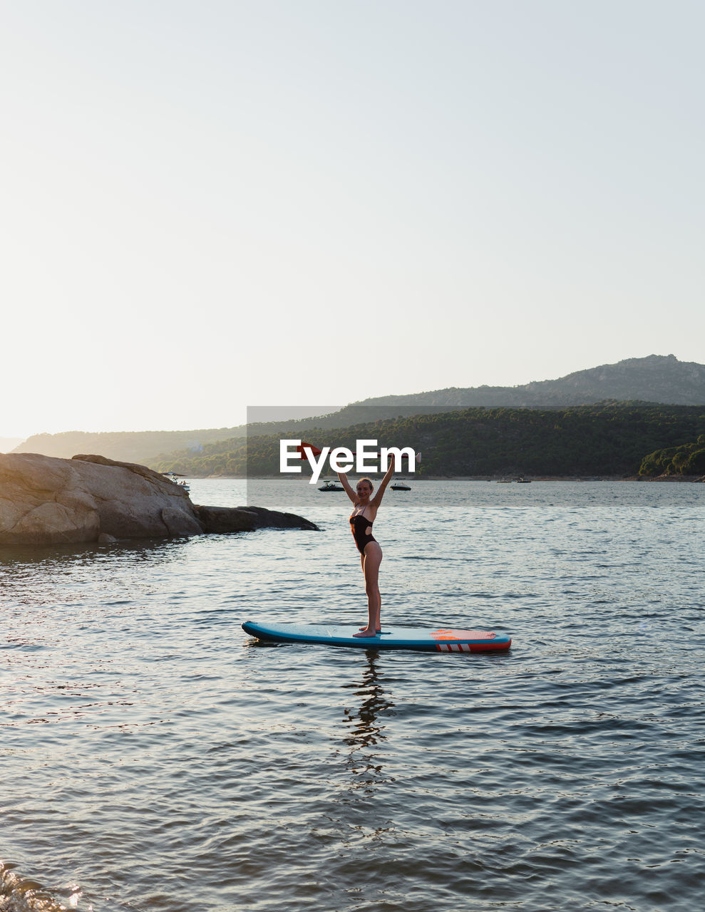 Full body of active female in swimwear floating on paddleboard on rippling lake during summer vacation in nature with forest