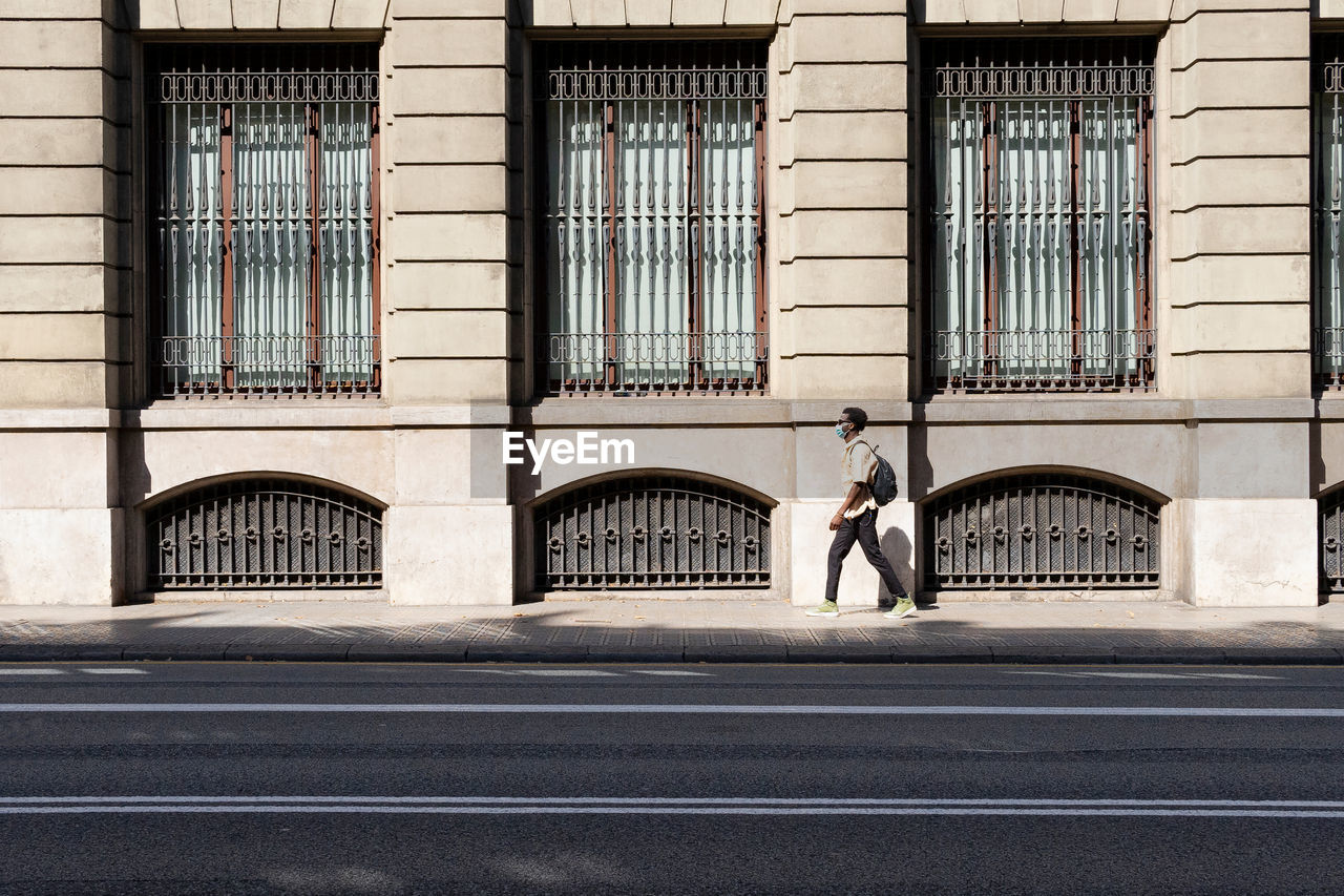 Side view of black male in medical mask walking along pavement near urban building on sunny day in barcelona during coronavirus epidemic