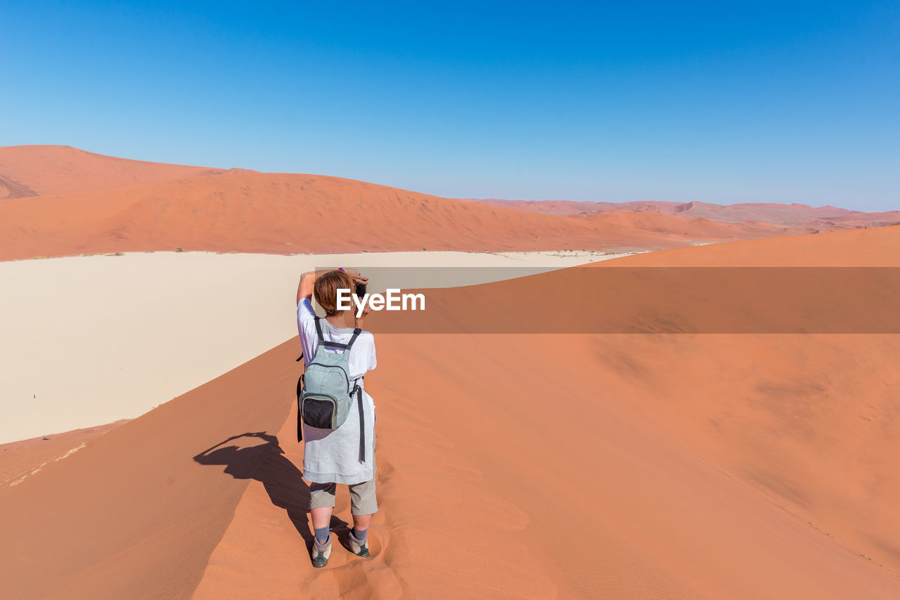 Rear view of woman photographing on desert against clear blue sky