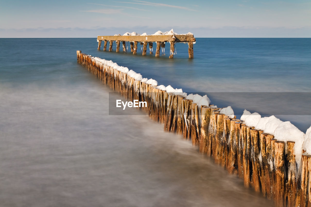 Snow on wooden posts in sea against sky