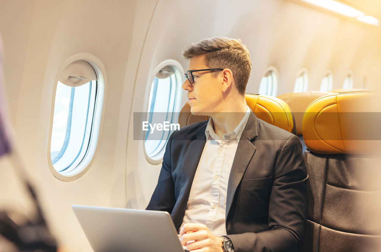 During a flight, a businessman in formal attire and glasses looks out the plane window