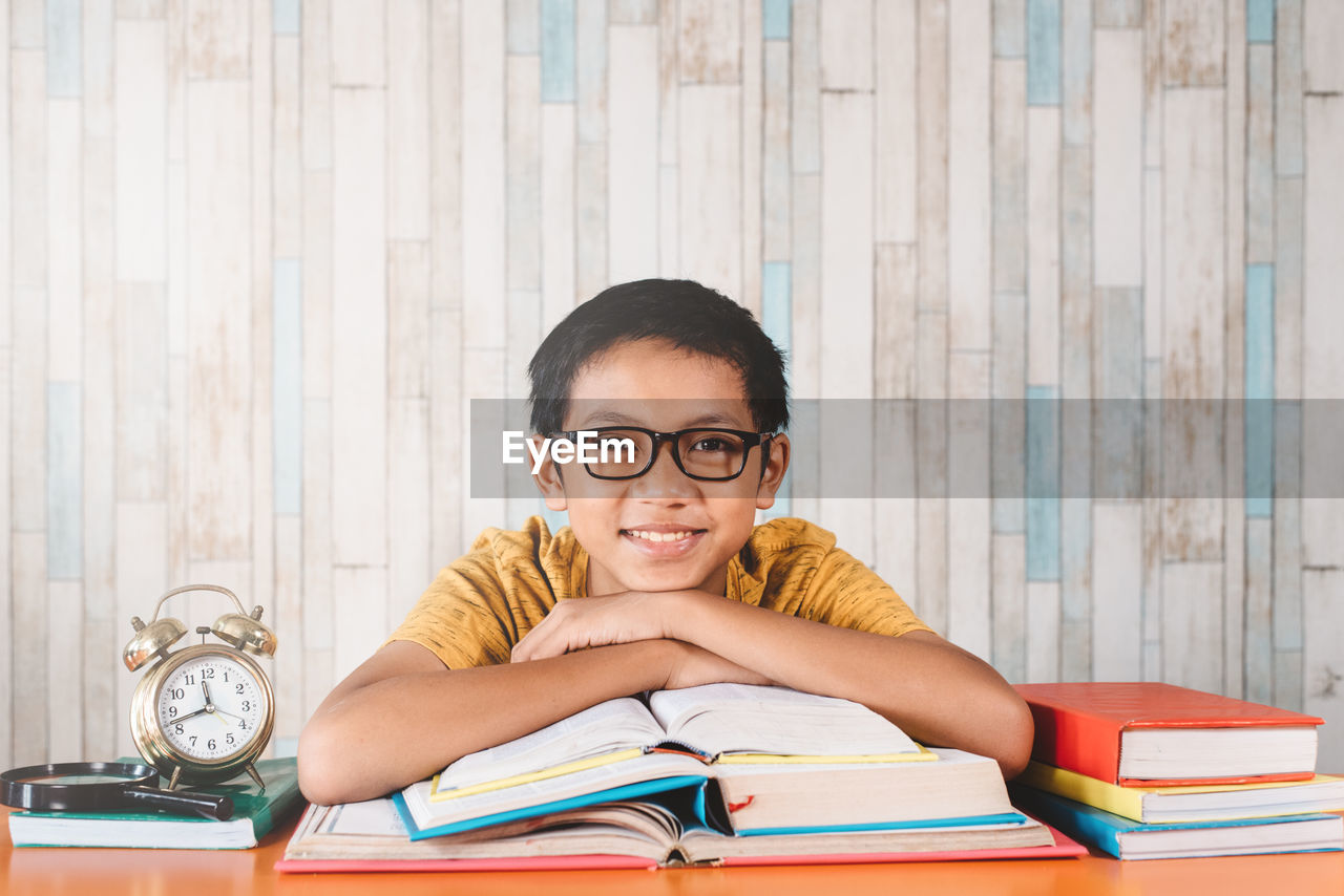 Young asian male student smiling while looking at camera sitting against table with books.