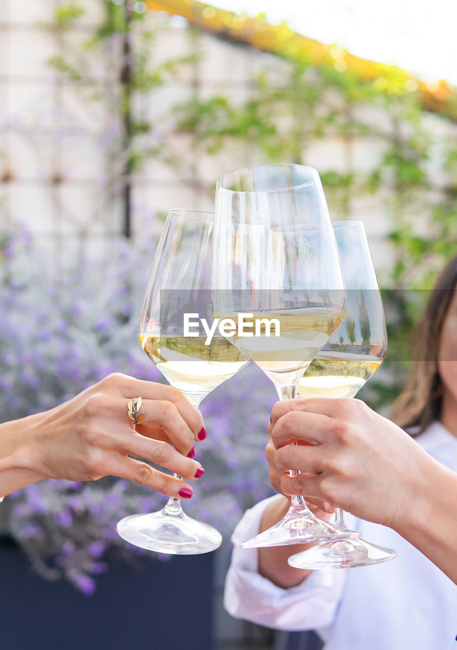 Cropped unrecognizable adult girlfriends in smart casual clothes sitting at restaurant table and clinking glasses of white wine during meeting on terrace