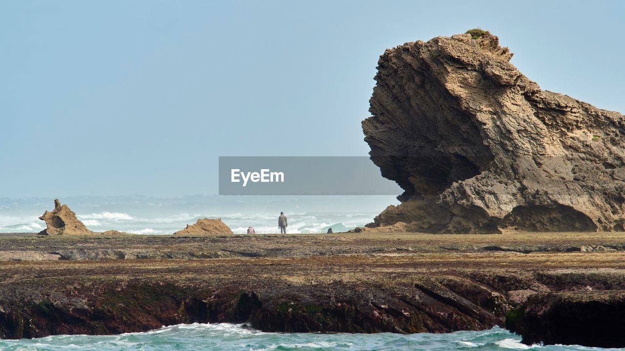 Figure dwarfed by rocks on sea shore against clear sky