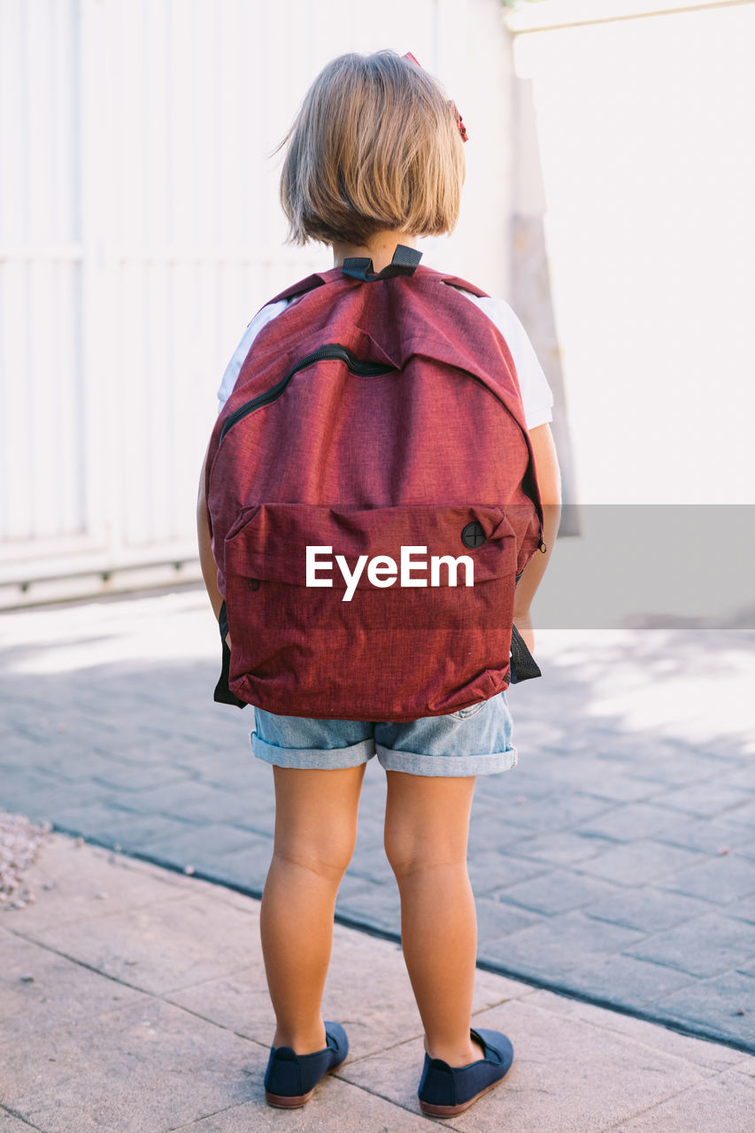 Back view of unrecognizable schoolchild with backpack standing on pavement in sunlight