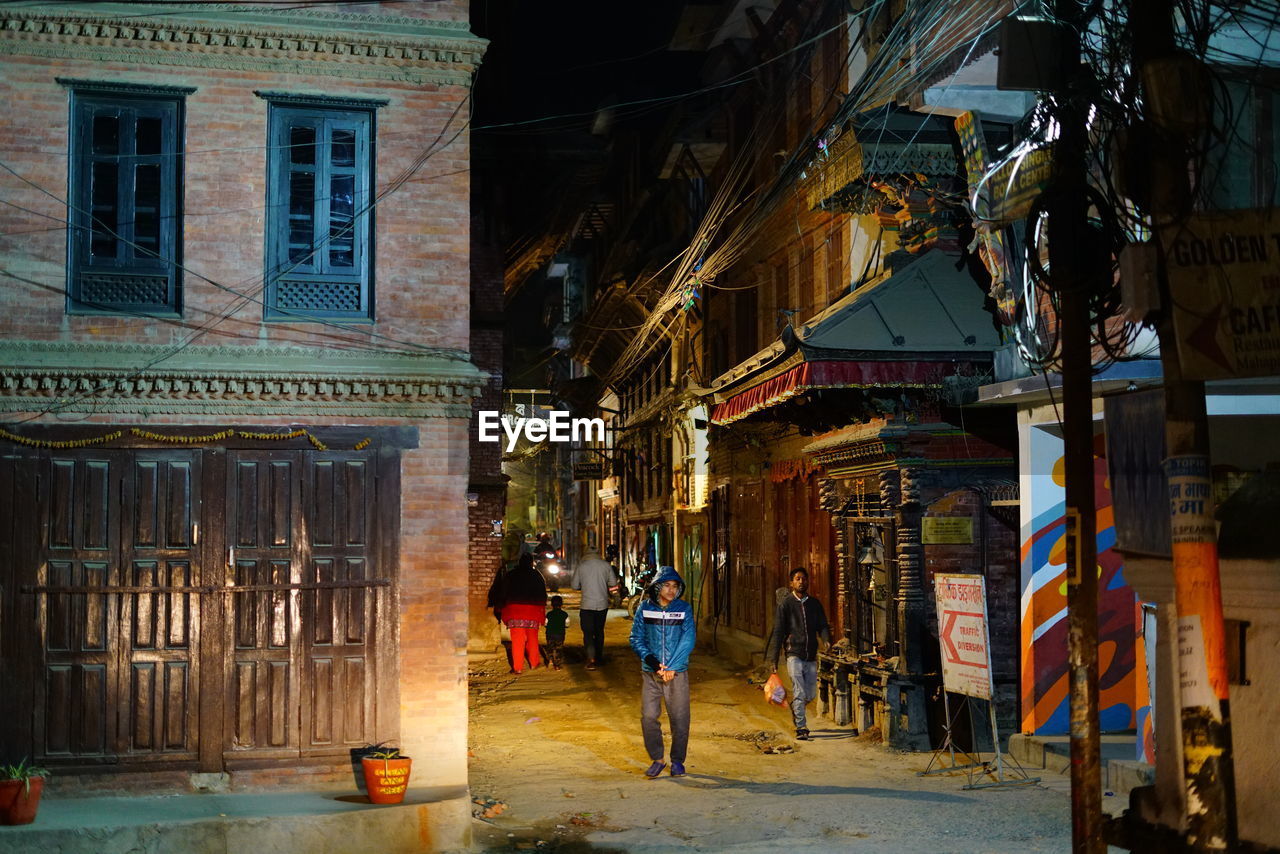 PEOPLE WALKING ON ILLUMINATED STREET AMIDST BUILDINGS IN CITY