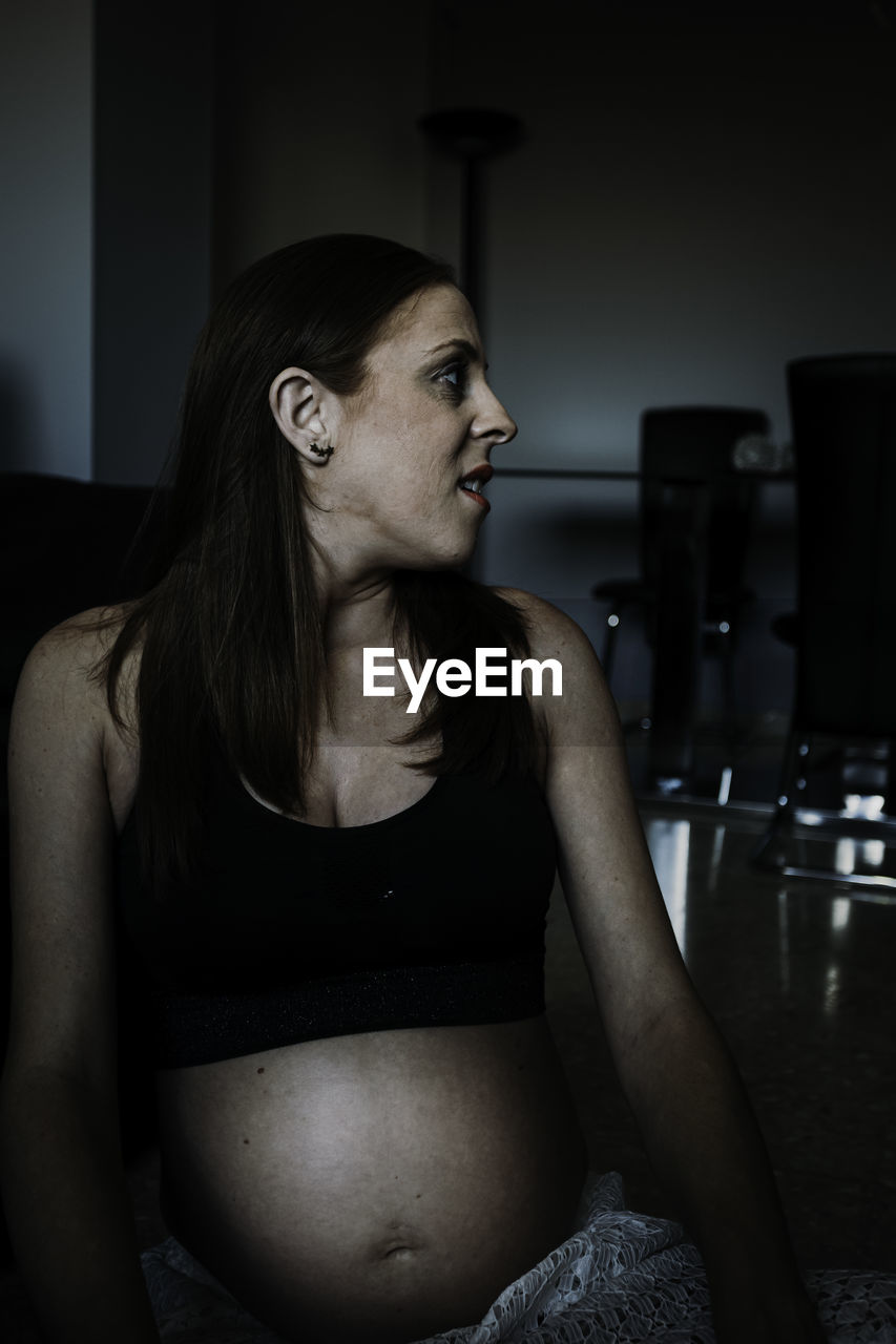 Young thoughtful pregnant lady in sportswear and sitting on floor in room