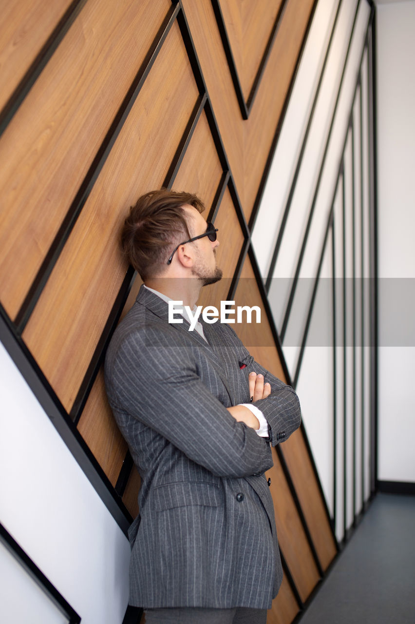A young man in a suit and sunglasses is standing near the wall with his hands folded