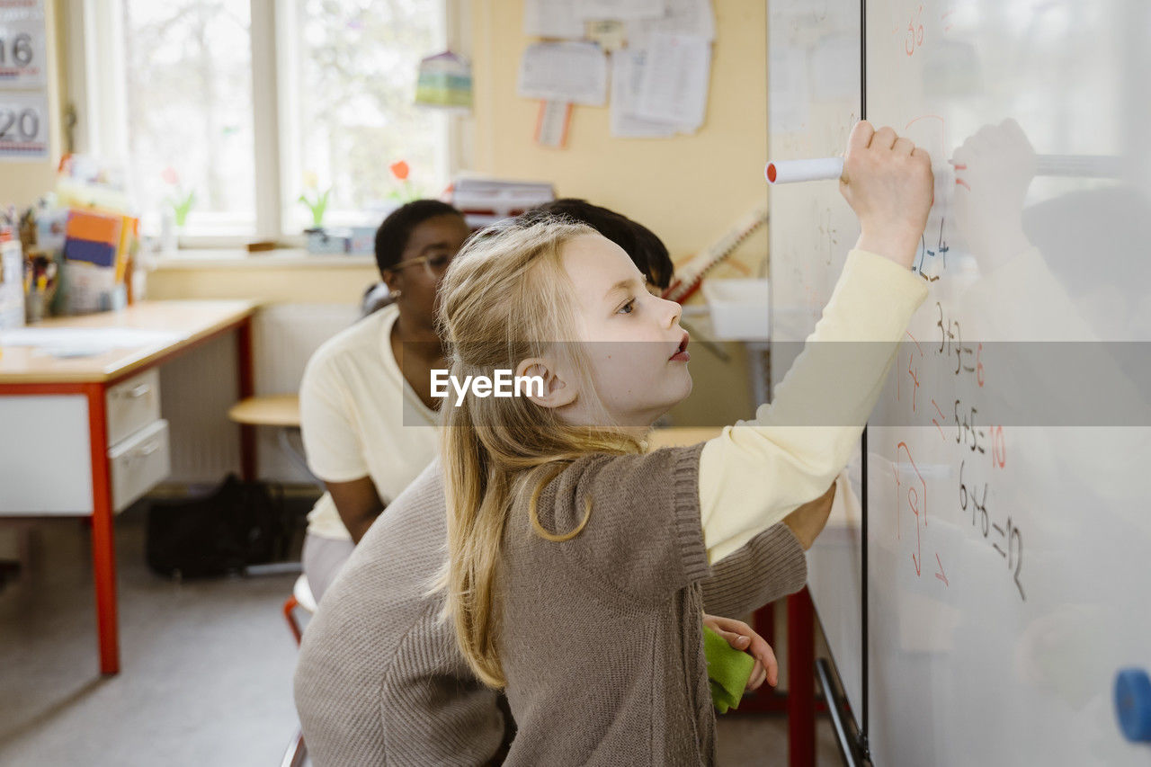 Blond girl solving maths problem while writing on whiteboard in classroom