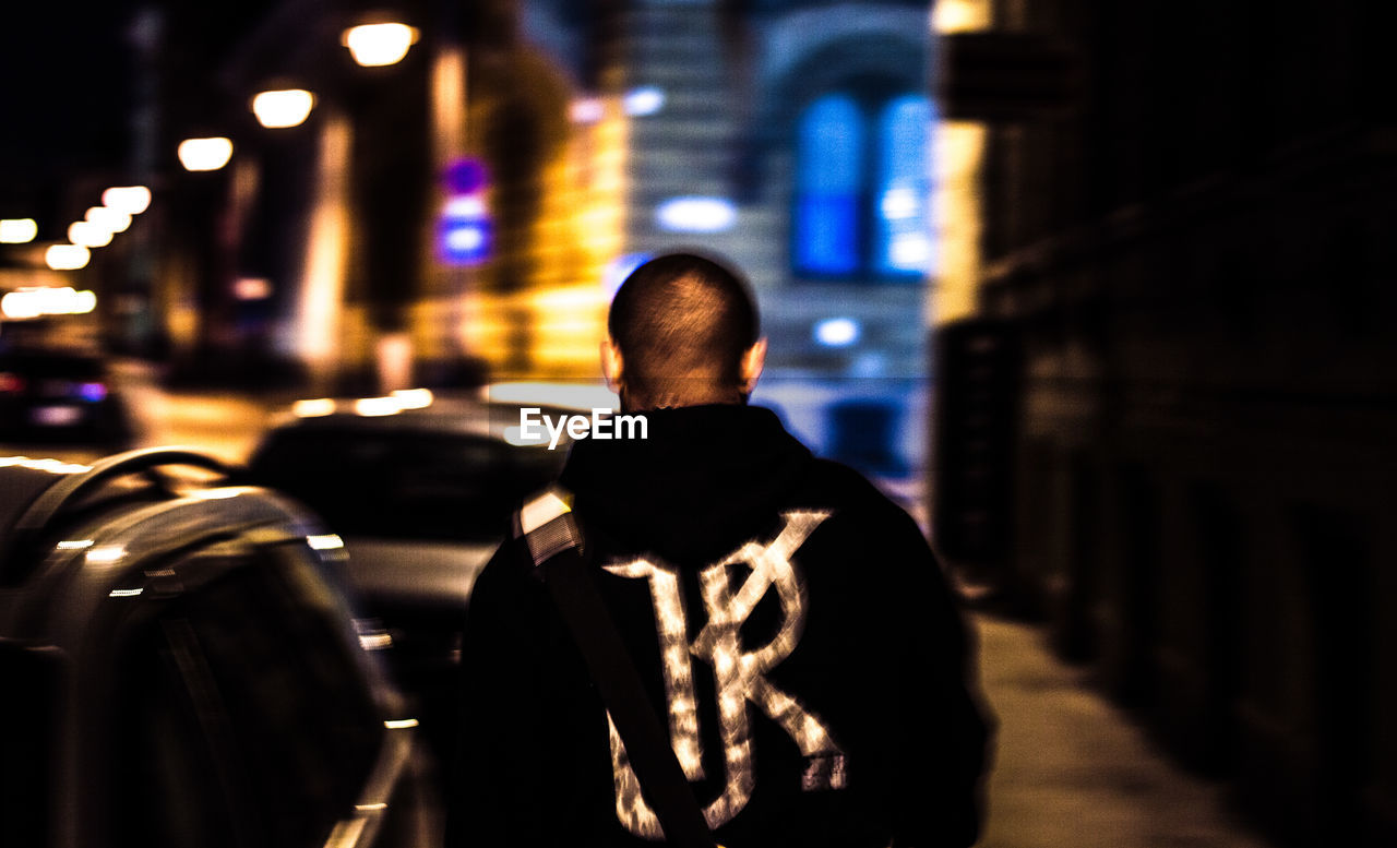 REAR VIEW OF MAN STANDING ON ILLUMINATED CITY AT NIGHT