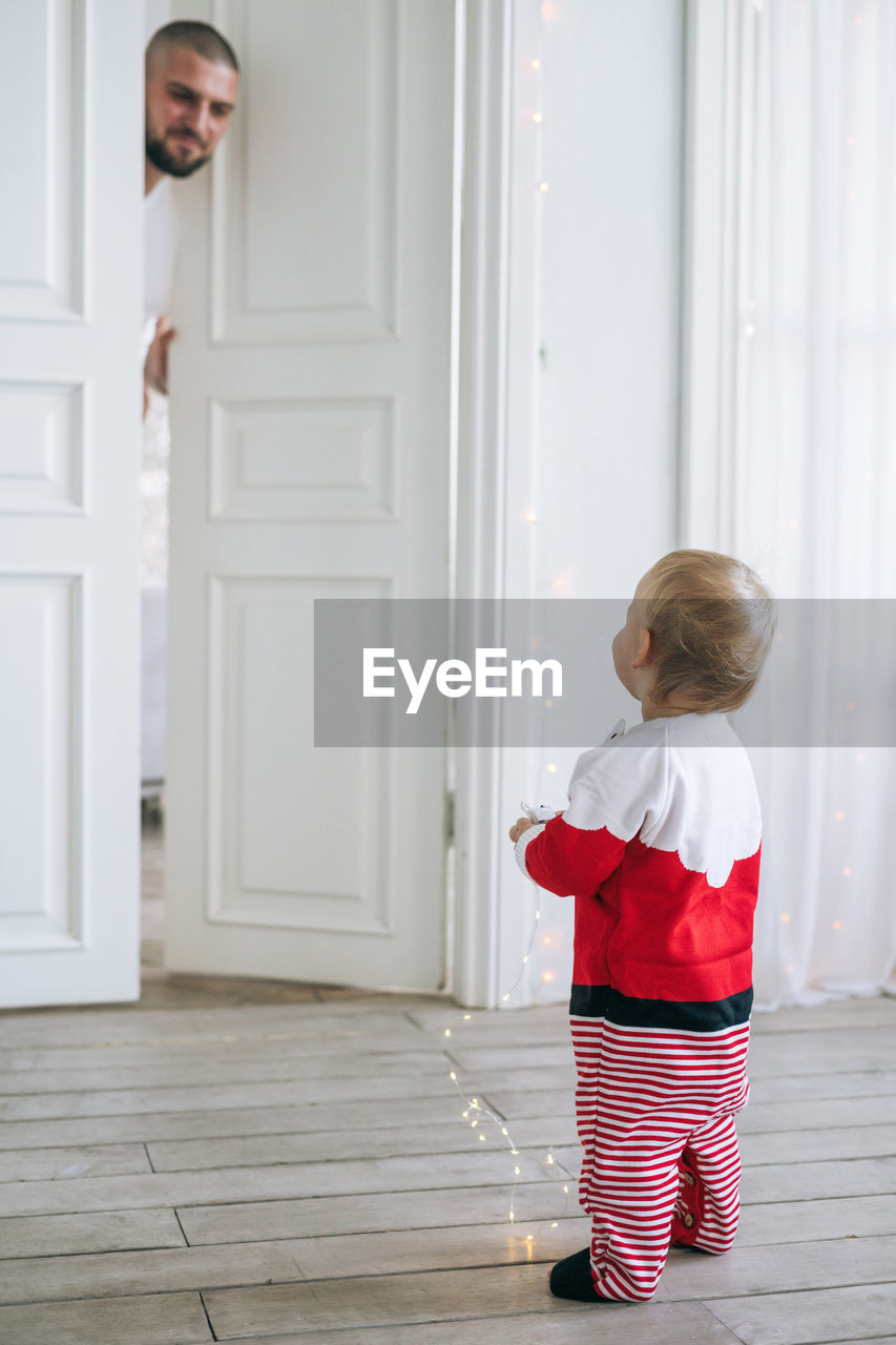 Cute little baby girl in santa costume standing in the large bright room looking at dad
