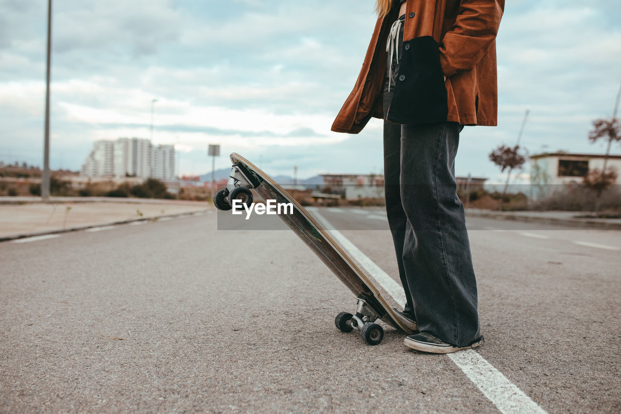 Side view of crop female skater in trendy outfit standing on asphalt road with cruiser skateboard in countryside