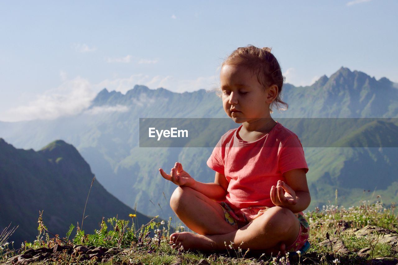 Girl meditating while sitting on mountain against sky