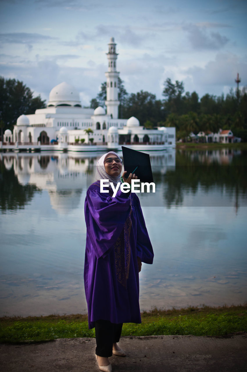 Woman holding mortarboard while standing against lake with mosque in background