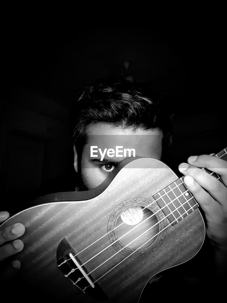 Portrait of young man covering face with ukulele against black background