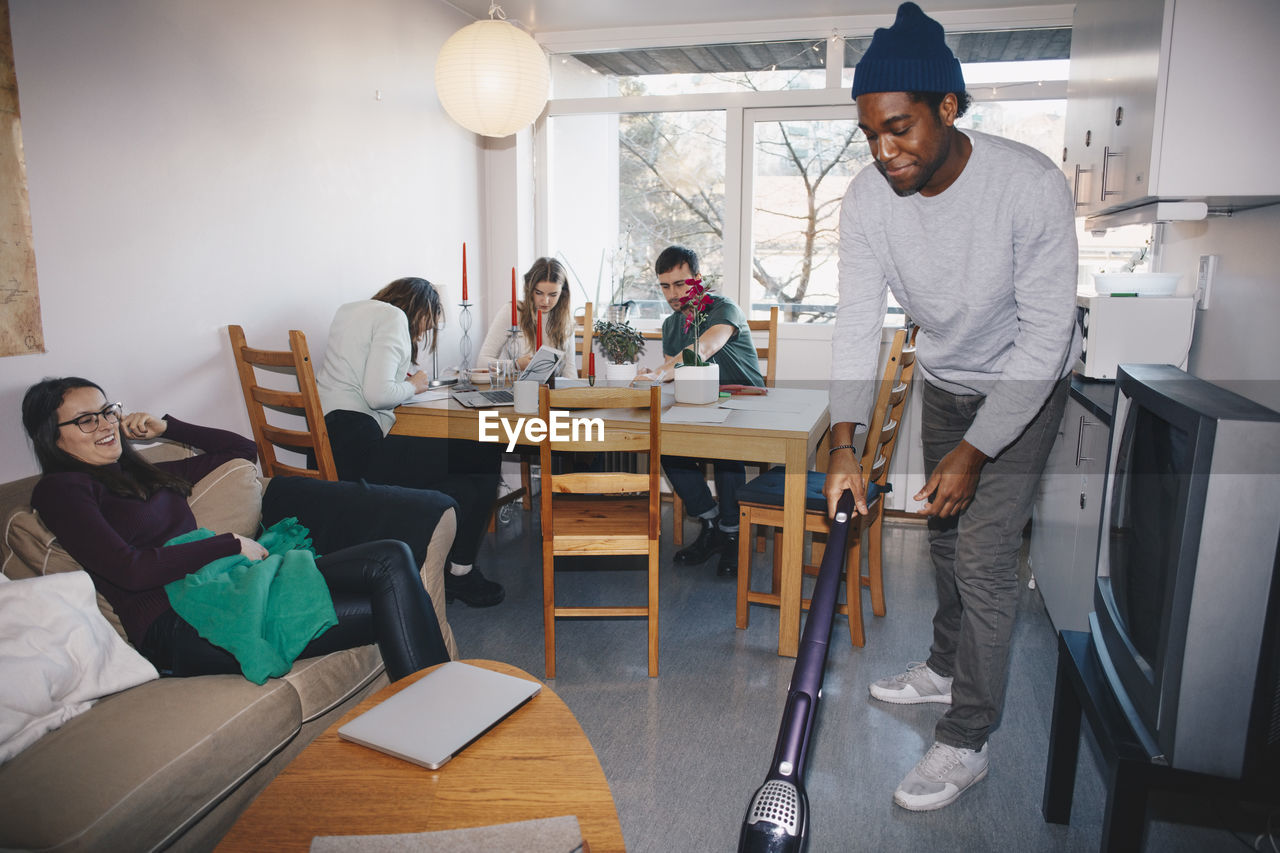 Man cleaning floor while friends sitting in college dorm room