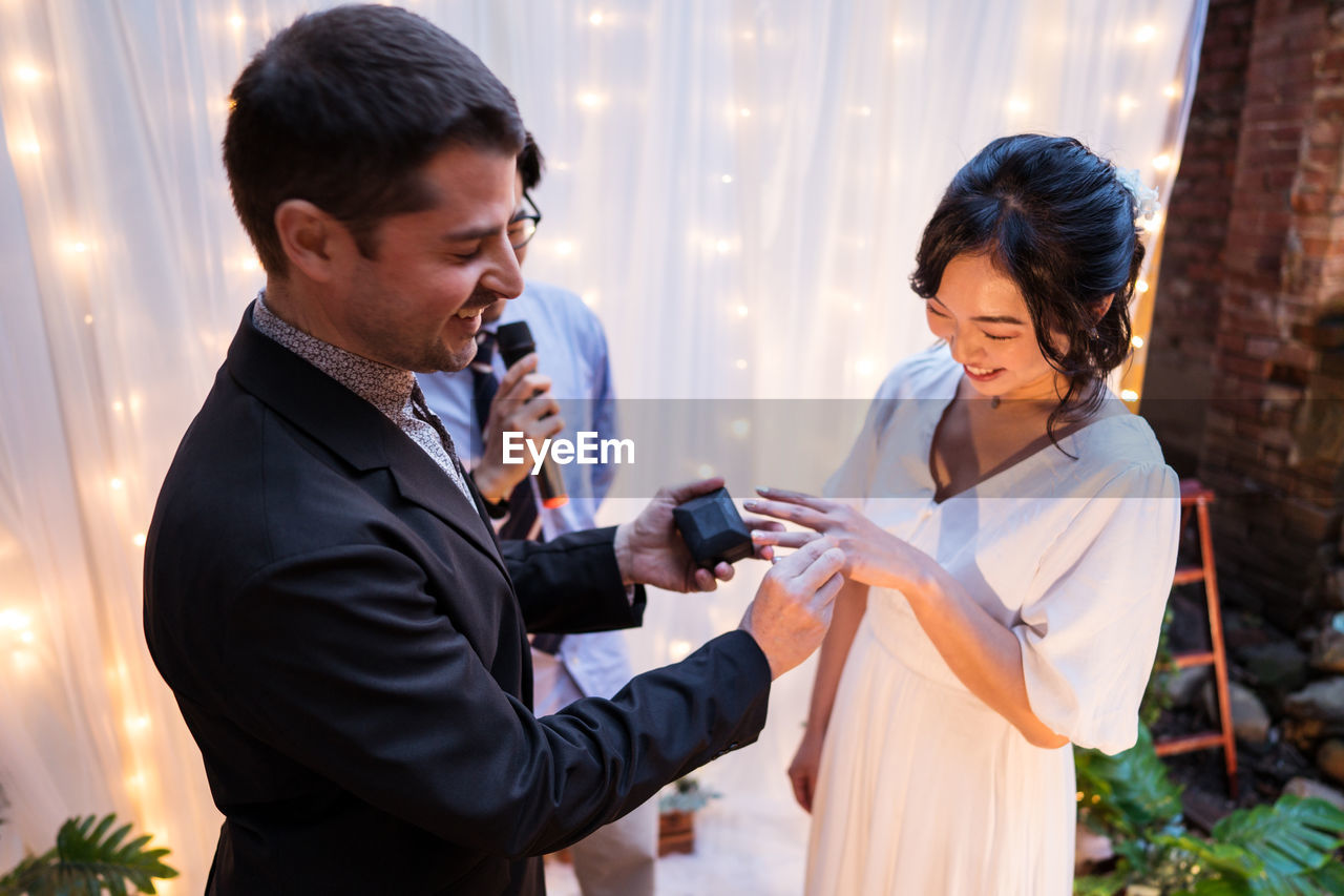 High angle of multiethnic newlywed couple smiling while standing near arch decorated with curtain and light bulb garlands while exchanging rings during wedding celebration