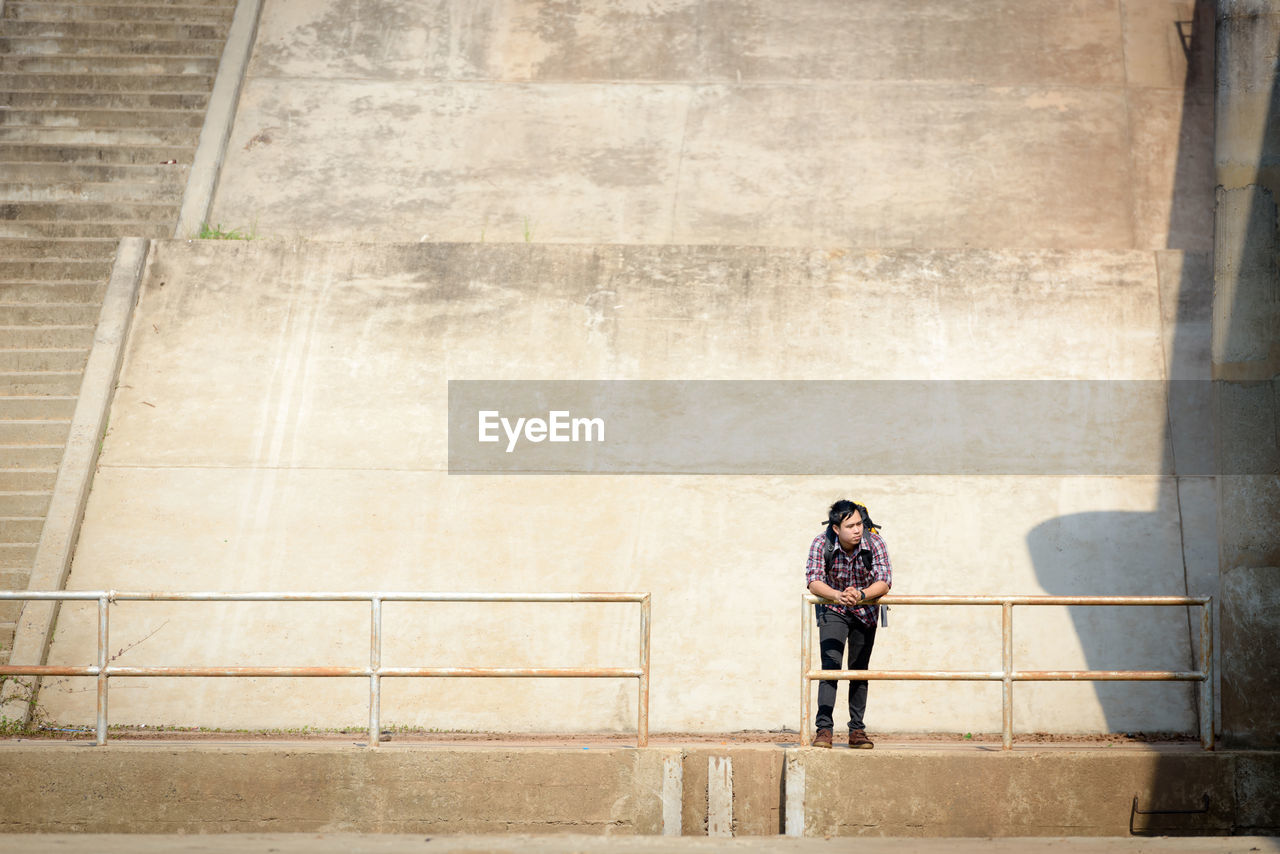 Full length of young man looking away while standing by railing against wall