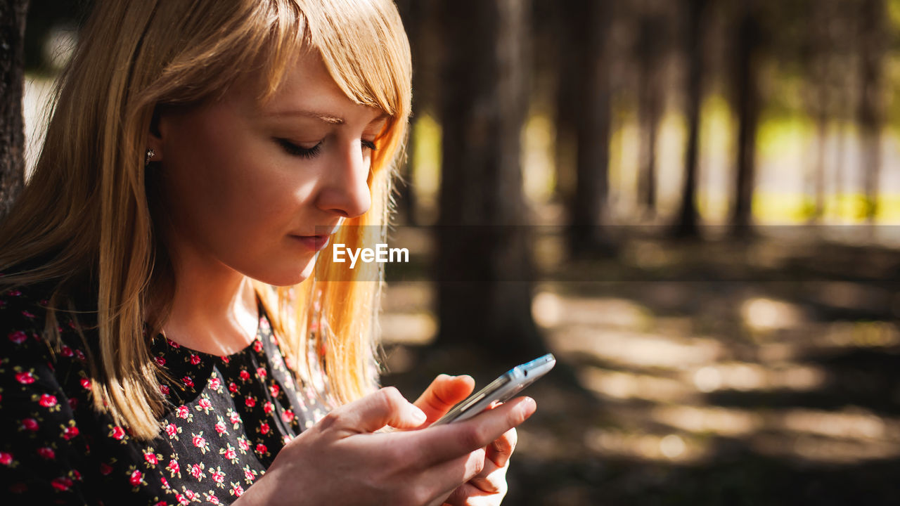 Woman using mobile phone while sitting by tree trunk in forest