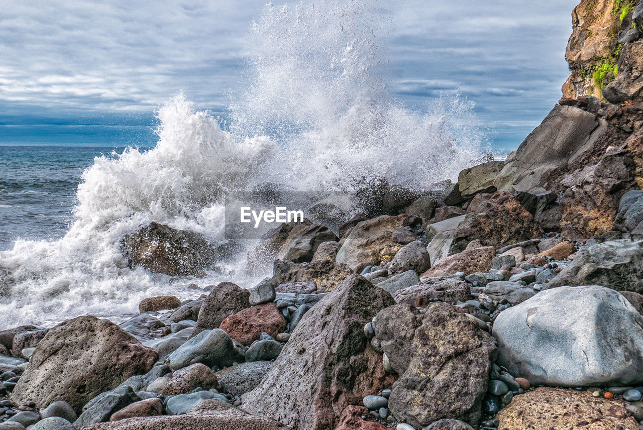 Waves splashing on rocks at shore against cloudy sky