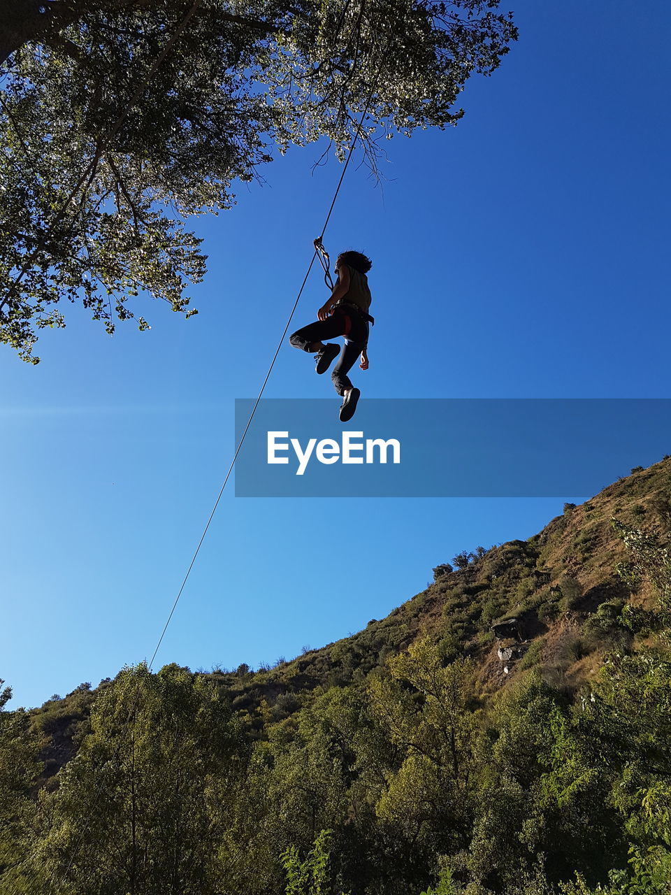 Low angle view of person hanging on zip line against clear blue sky