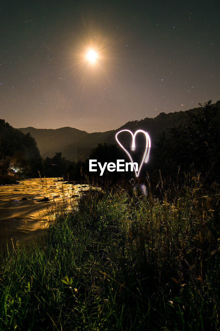 Heart shape light painting over grassy field by river at night