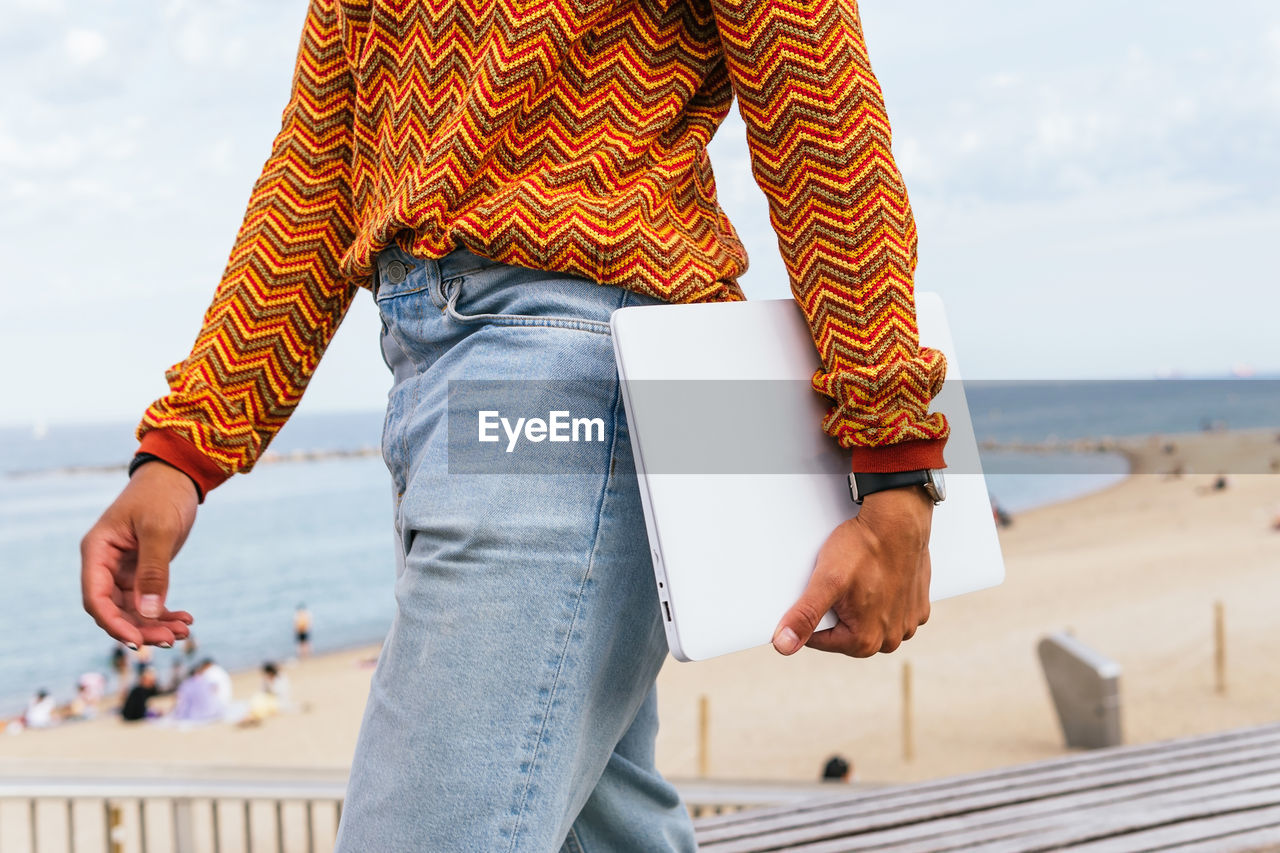 Side view of crop anonymous ethnic male in stylish colorful striped shirt and jeans carrying laptop while walking on embankment near sandy beach and sea