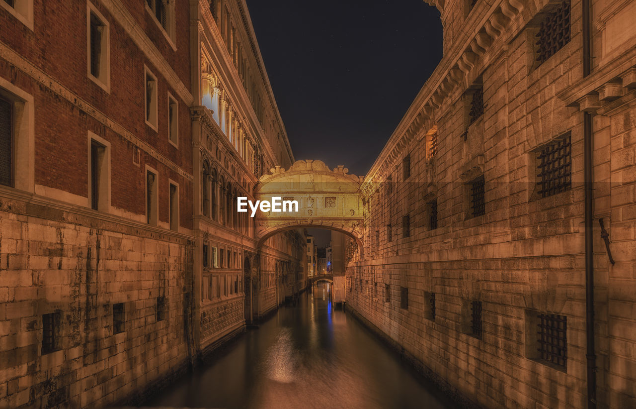 PEOPLE WALKING ON ILLUMINATED STREET AMIDST BUILDINGS IN CITY