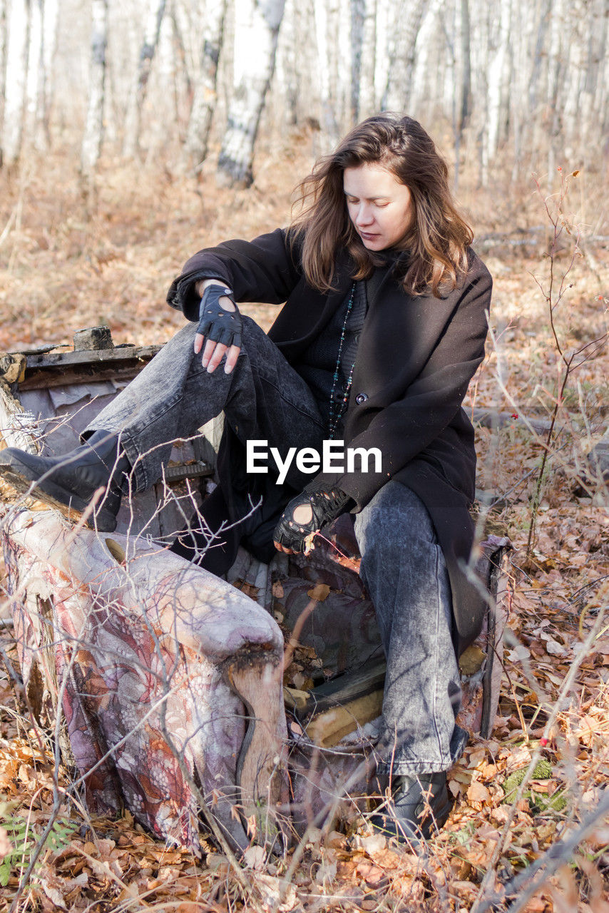 A young woman in casual attire sits on an old suitcase in a serene forest. 