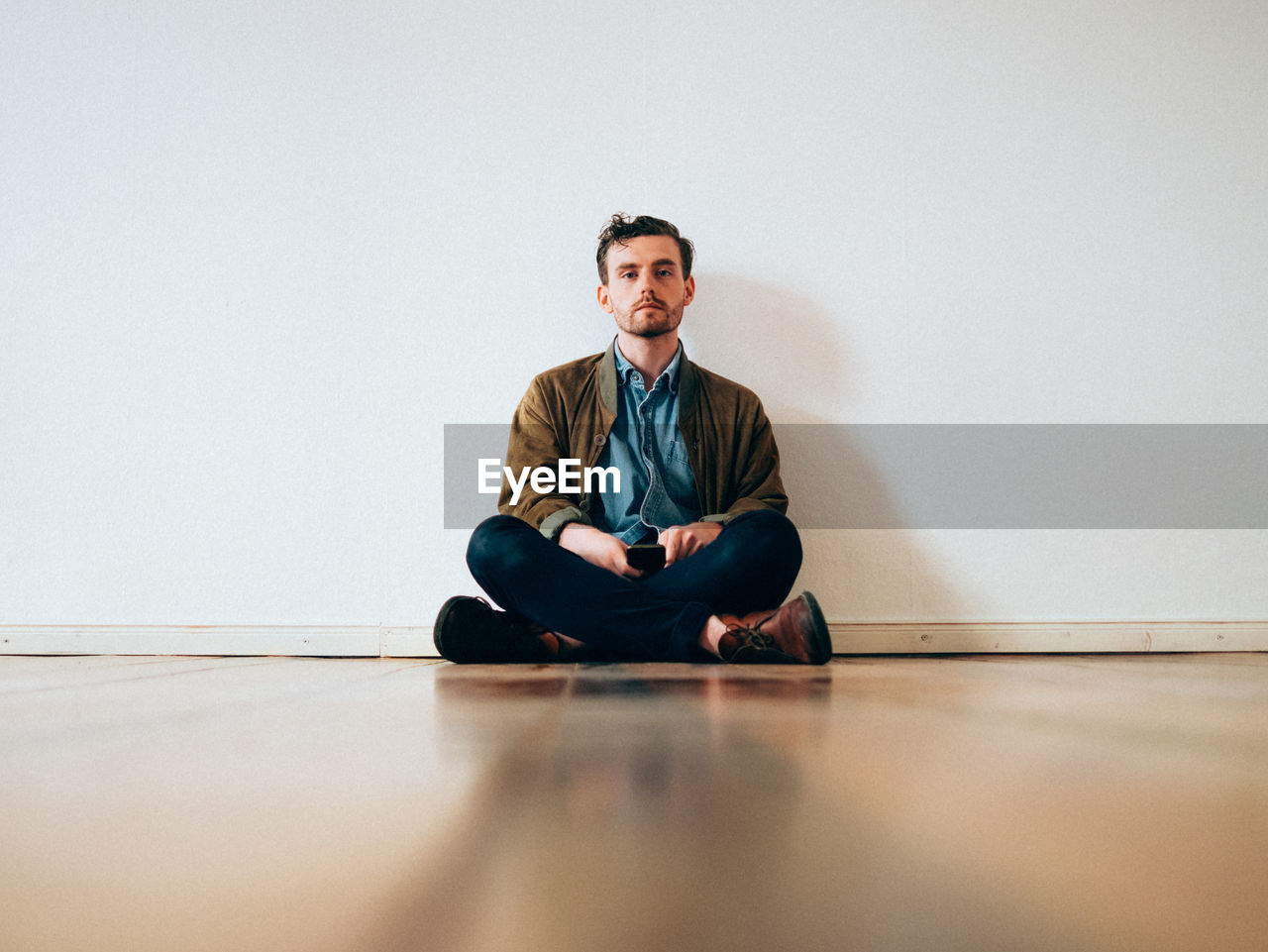 Full length portrait of serious man sitting on hardwood floor against white wall