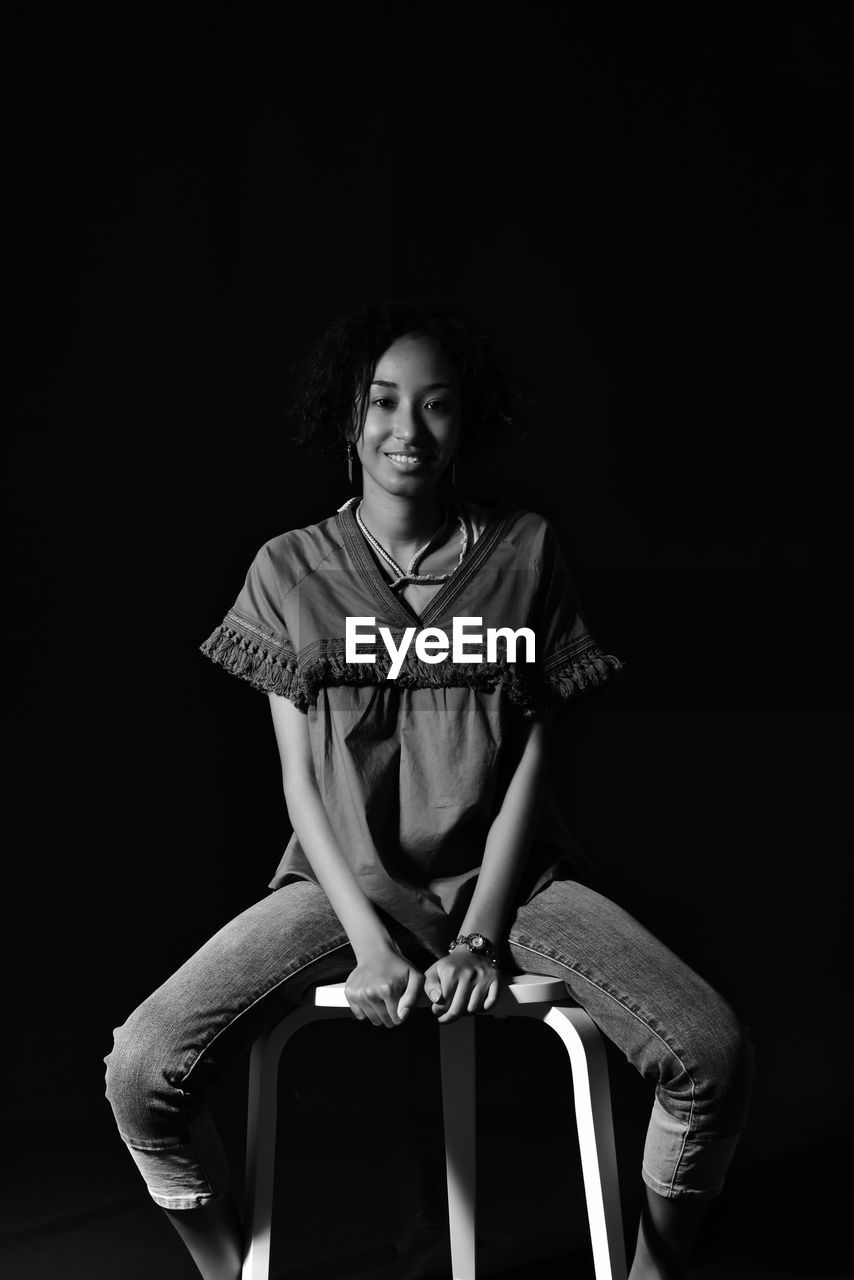 Portrait of young woman sitting on chair against black background