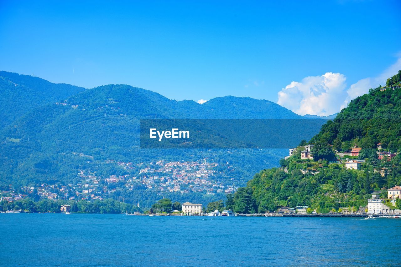 Scenic view of town by sea and mountains against blue sky